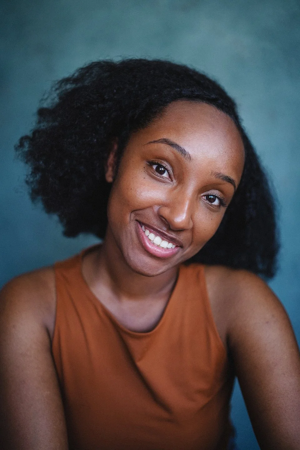 A smiling woman with natural curly hair and wearing a sleeveless brown top poses against a solid blue background.