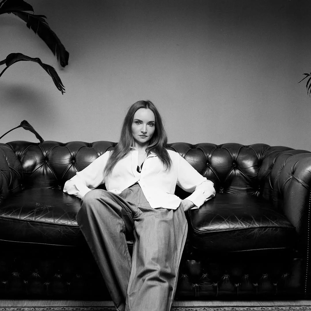 A woman with long hair sitting on a leather sofa, wearing a white blouse and wide trousers, in a black and white photograph.