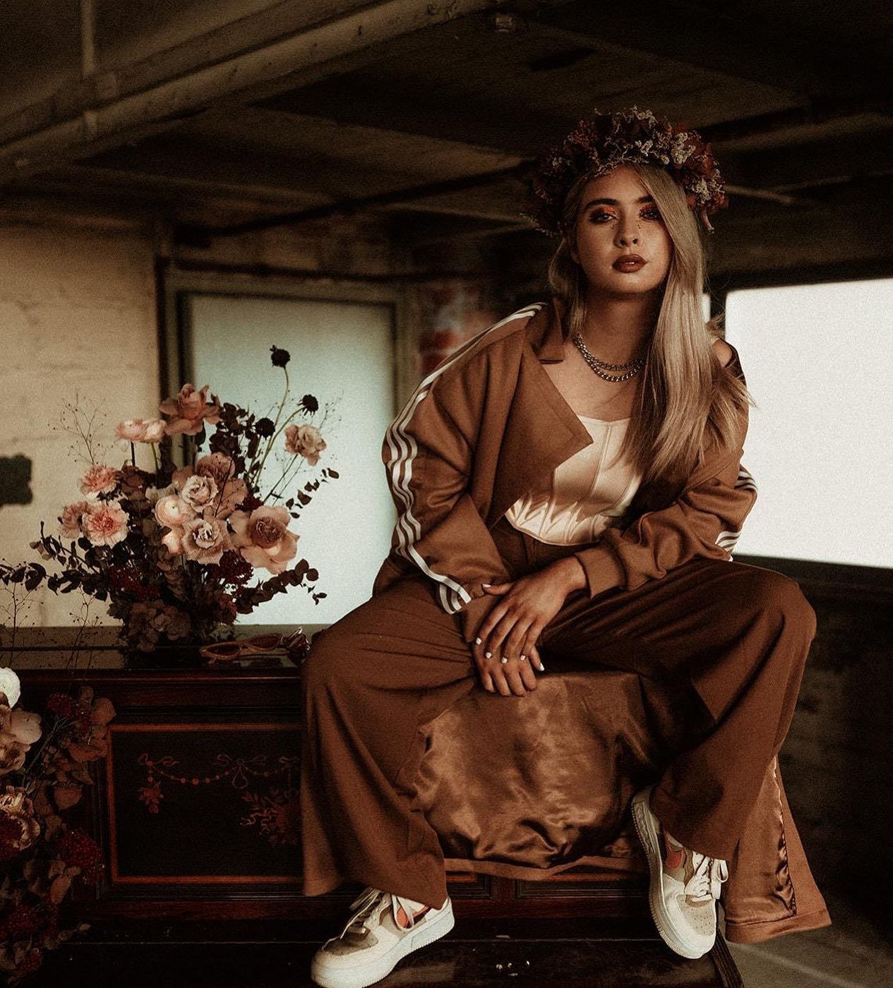 A woman with long blonde hair wearing a flower crown, a white top, brown track suit, and white sneakers, sitting on a wooden surface beside a floral arrangement in a rustic interior.