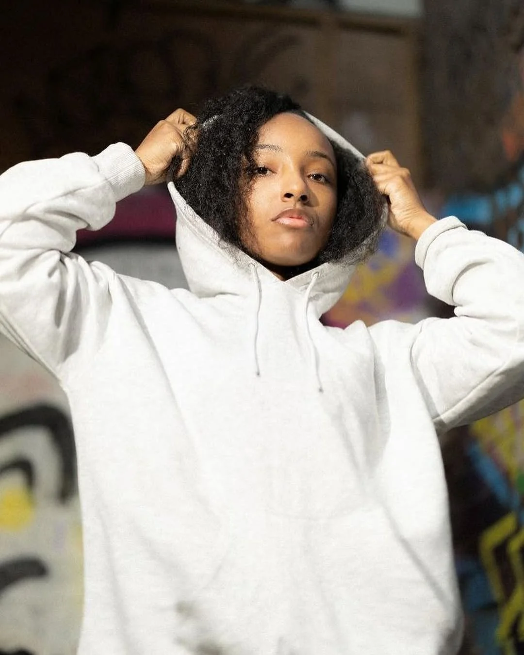 Young woman adjusting the hood of her white hoodie in front of colorful graffiti art.