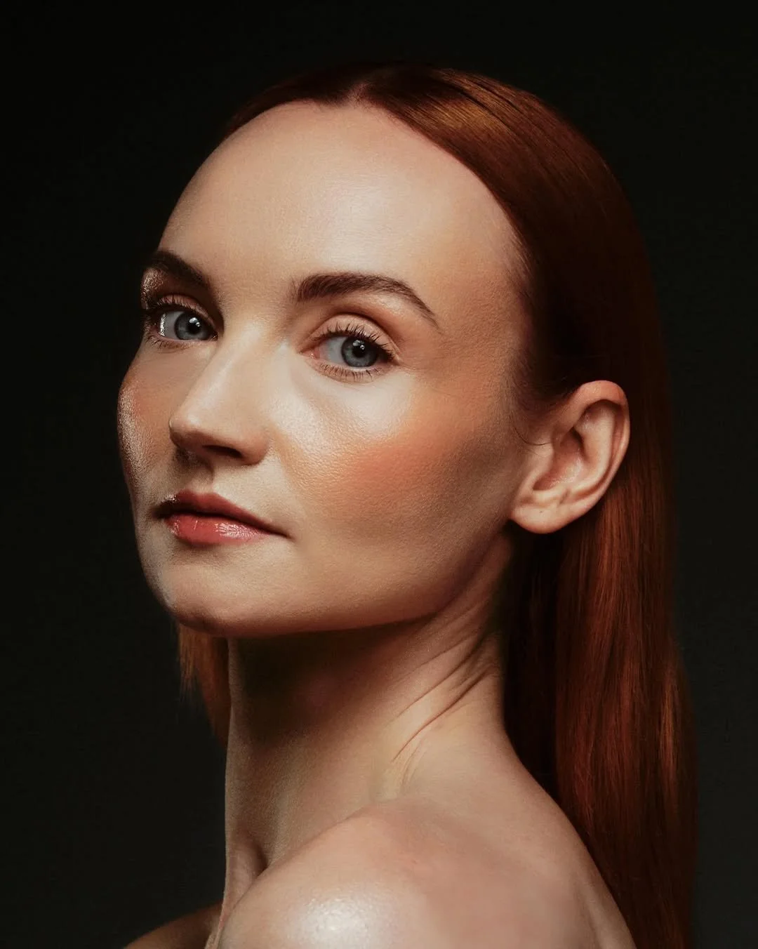 Close-up portrait of a woman with red hair, blue eyes, natural makeup, and fair skin against a dark background.