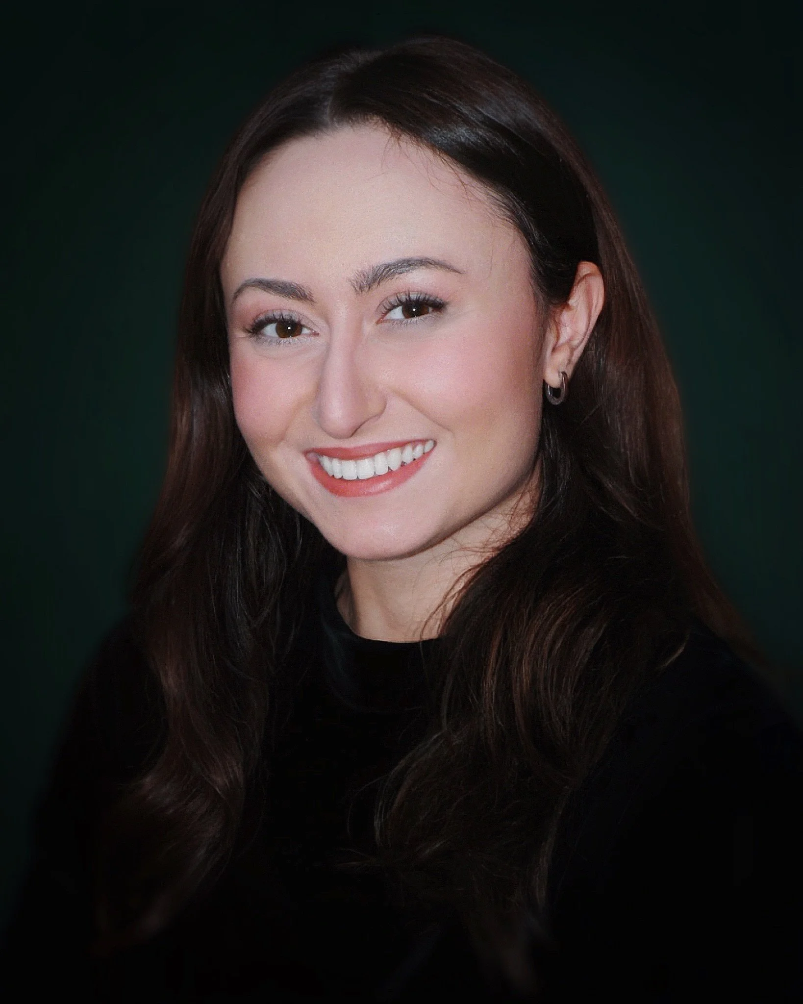 A woman with long, dark brown hair smiling and showing her teeth, against a dark background.