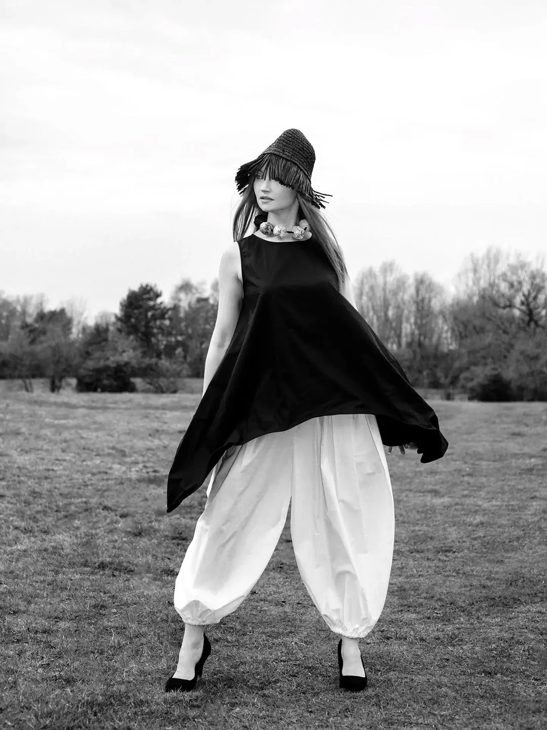 A woman standing outdoors in a grassy field, wearing a wide-brimmed hat, a sleeveless black top, white balloon pants, and high heels, with trees in the background.