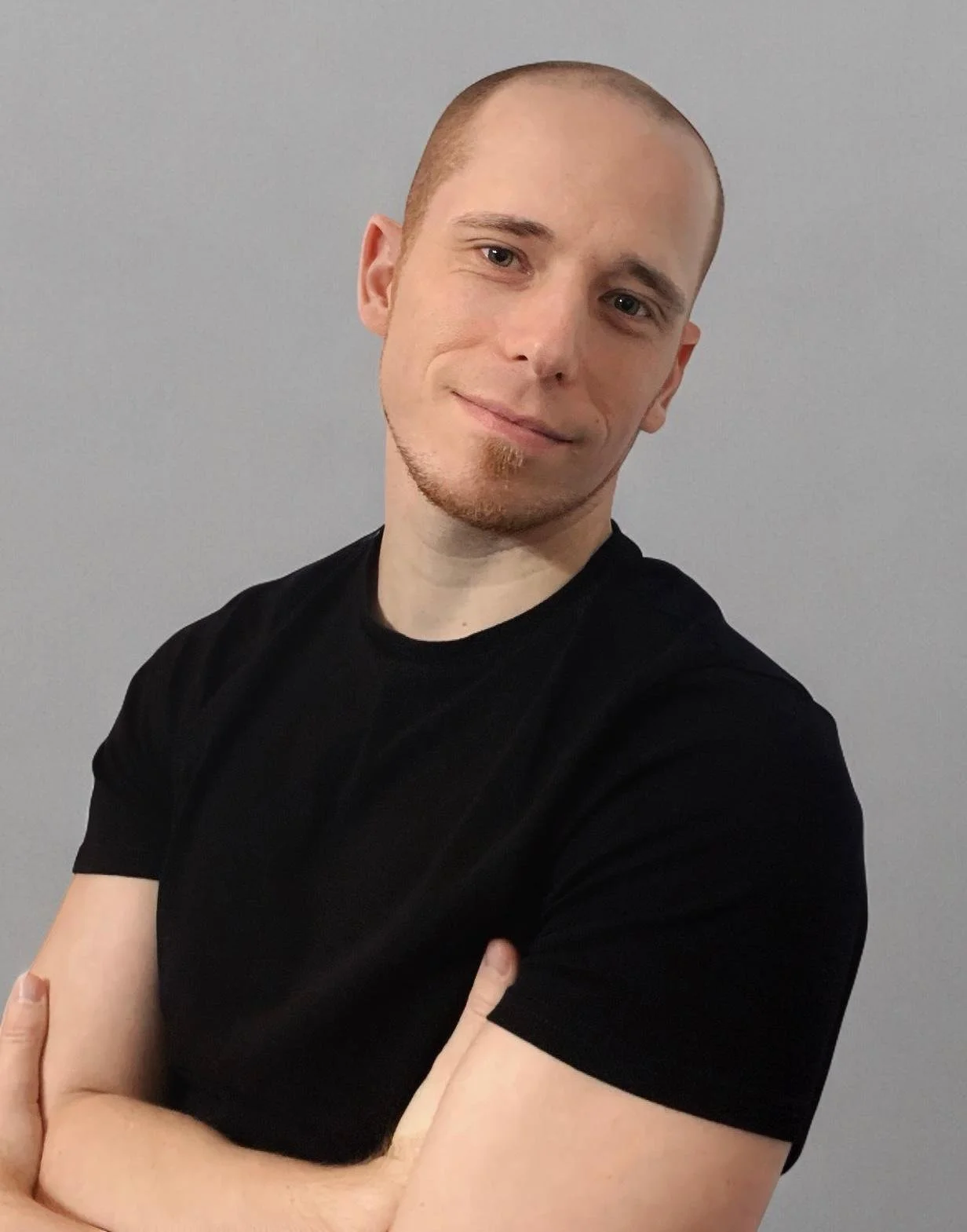 A man with a shaved head and a small goatee, wearing a black t-shirt, stands with crossed arms and a slight smile against a plain gray background.