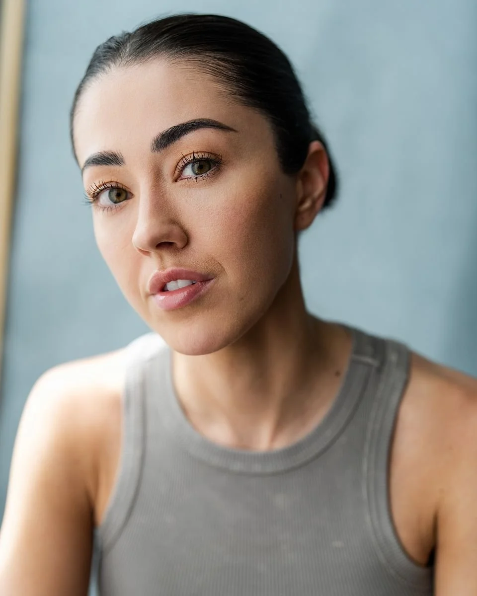 Close-up of a young woman with dark hair pulled back, wearing a gray sleeveless top, looking at the camera with a neutral expression.