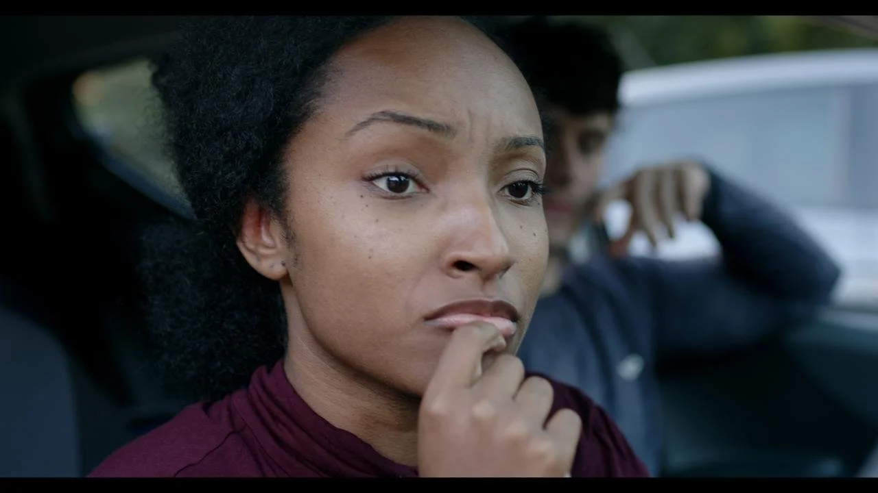 Close-up of a concerned woman with natural hair sitting in a car, with a teenage boy in the background making a gesture as if talking on the phone.