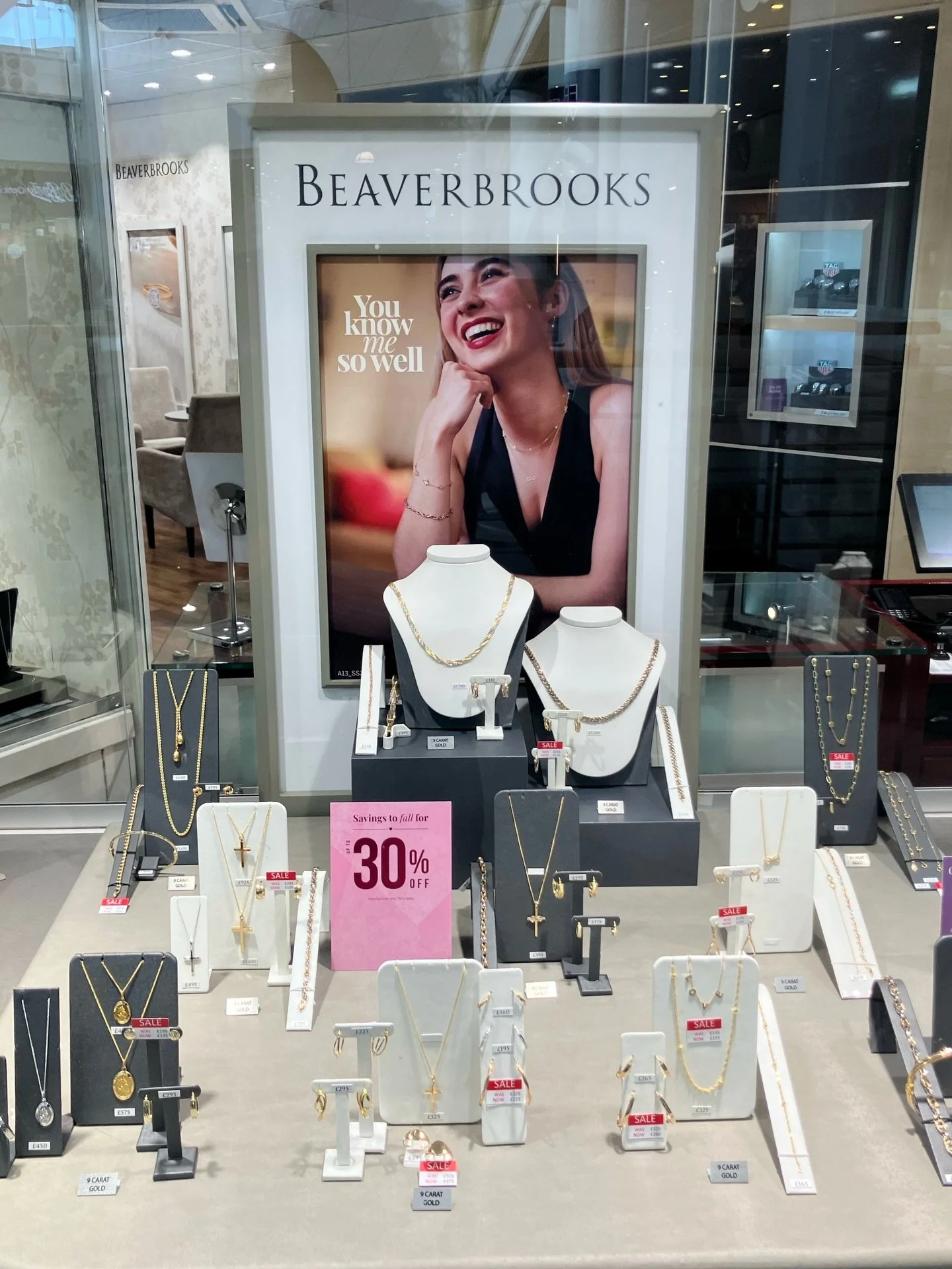 Display of gold jewelry including necklaces, earrings, and rings with sale tags, in a jewelry store window with a large poster of a smiling woman and a sign indicating 30% off.