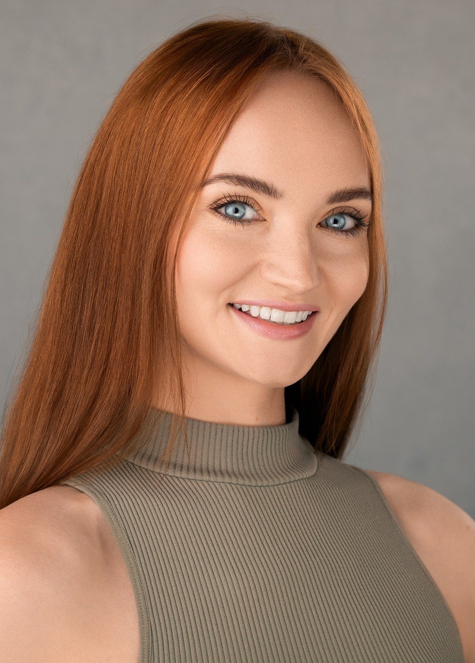 Close-up of a young woman with long red hair and blue eyes, smiling, wearing a sleeveless beige top, against a neutral gray background.