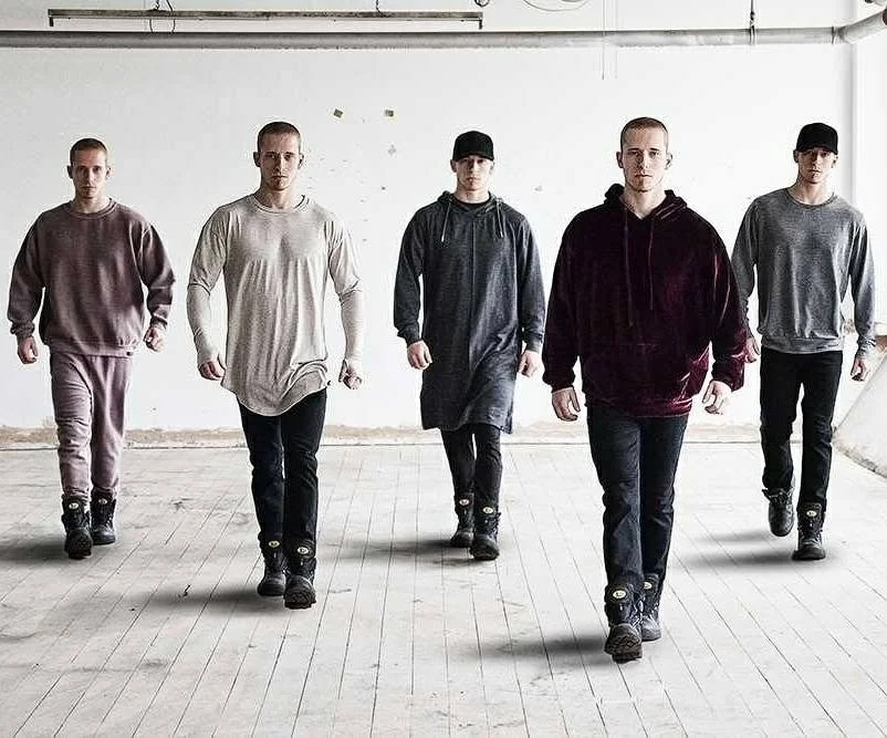 Five young men walking towards the camera in an empty room with white walls and wooden floor.