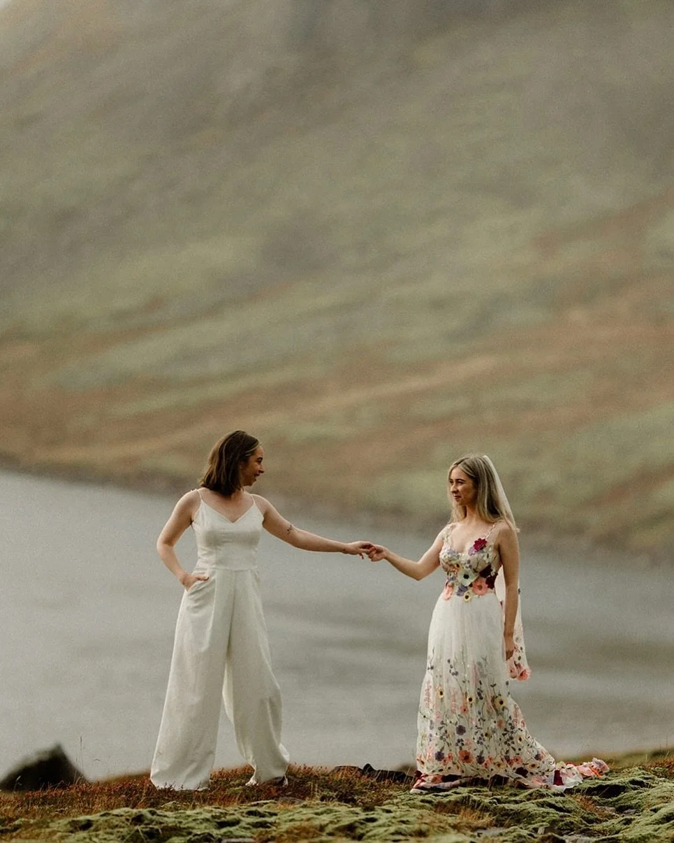 Two women holding hands on a grassy terrain with a large hill in the background.