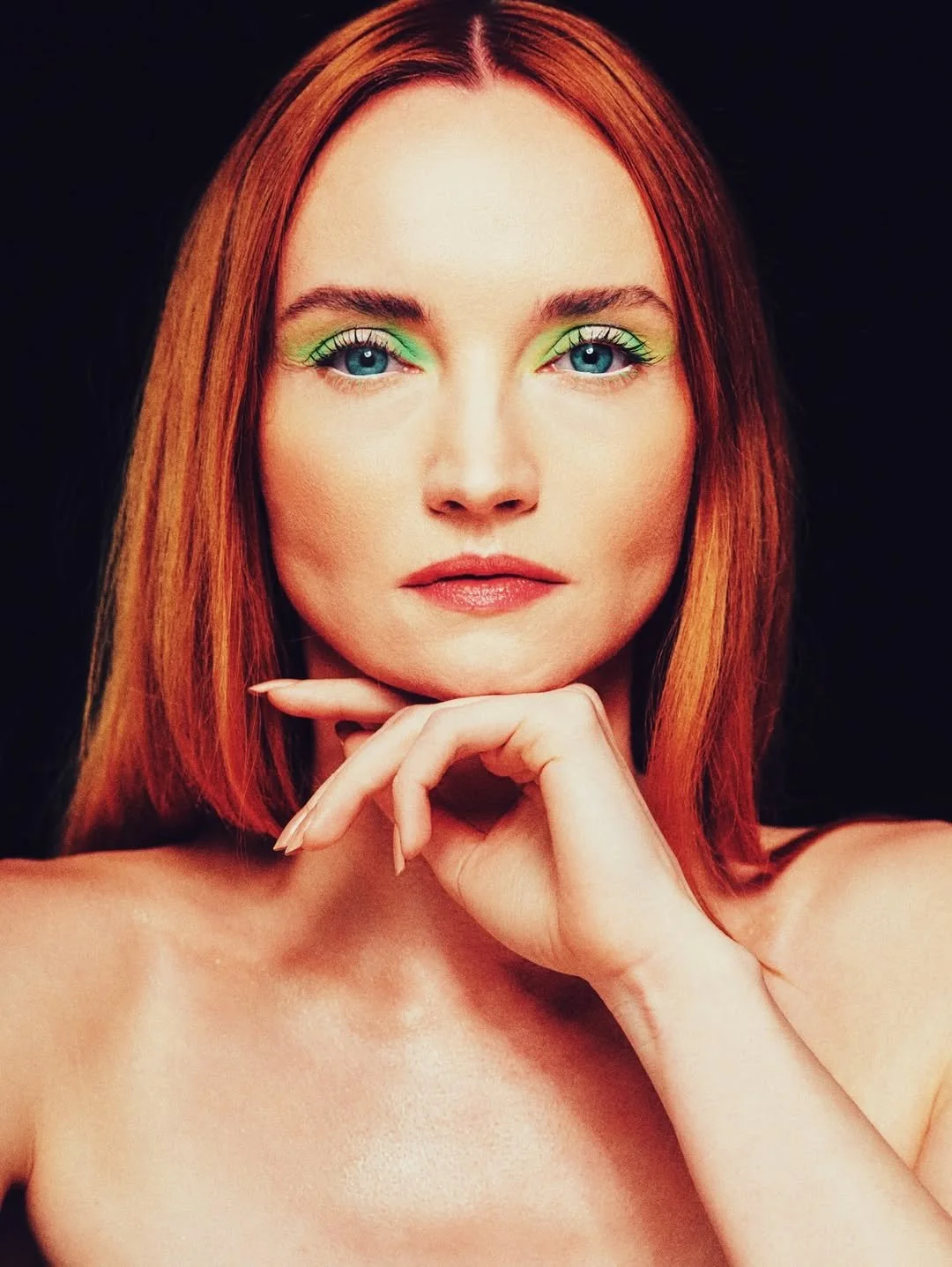 A woman with red hair looking directly at the camera against a black background, with her hand resting on her chin.