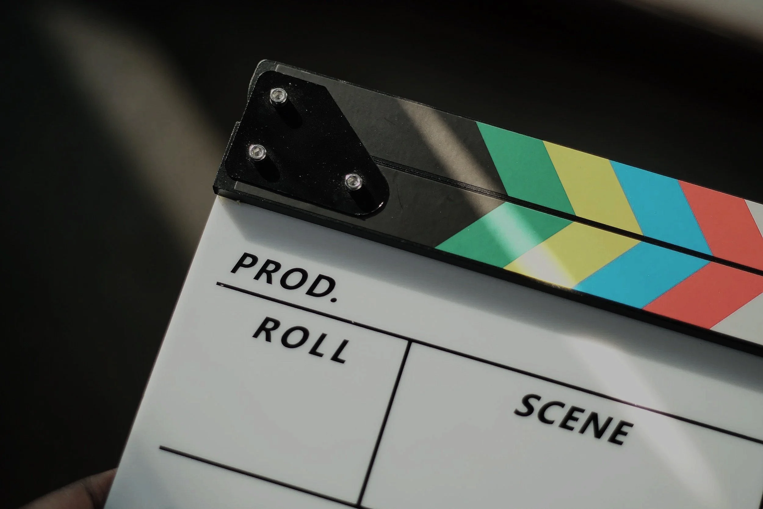Close-up of a film clapperboard with colorful diagonal stripes and black screws, with part of a white sheet that has black text and a grid for scene, take, roll, and scene number.