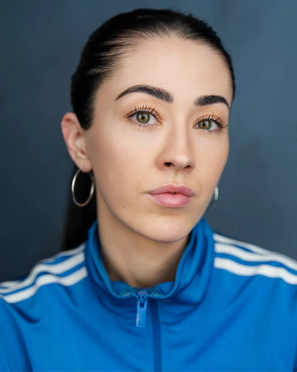 Close-up portrait of a young woman with dark hair pulled back, wearing silver hoop earrings and a blue sports jacket with white stripes, against a dark background.
