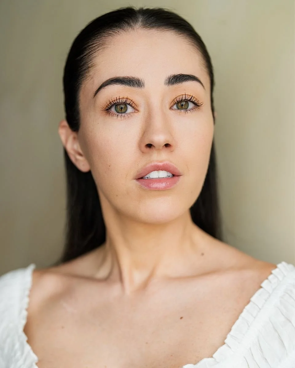 Close-up portrait of a young woman with dark hair, fair skin, and green eyes, wearing a white top with ruffled edges, against a neutral background.