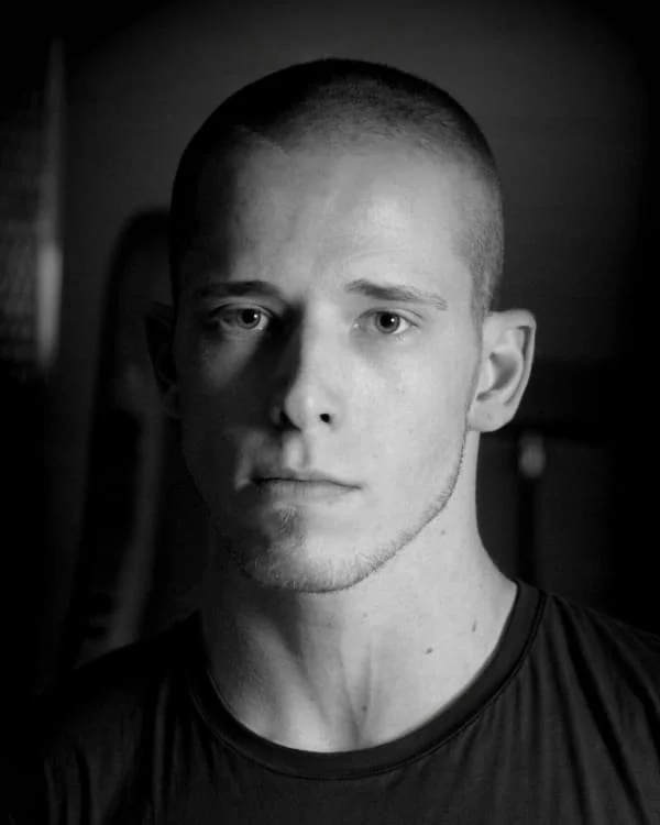 Black and white portrait of a young man with a buzz cut, serious expression, wearing a dark shirt, looking directly at the camera.