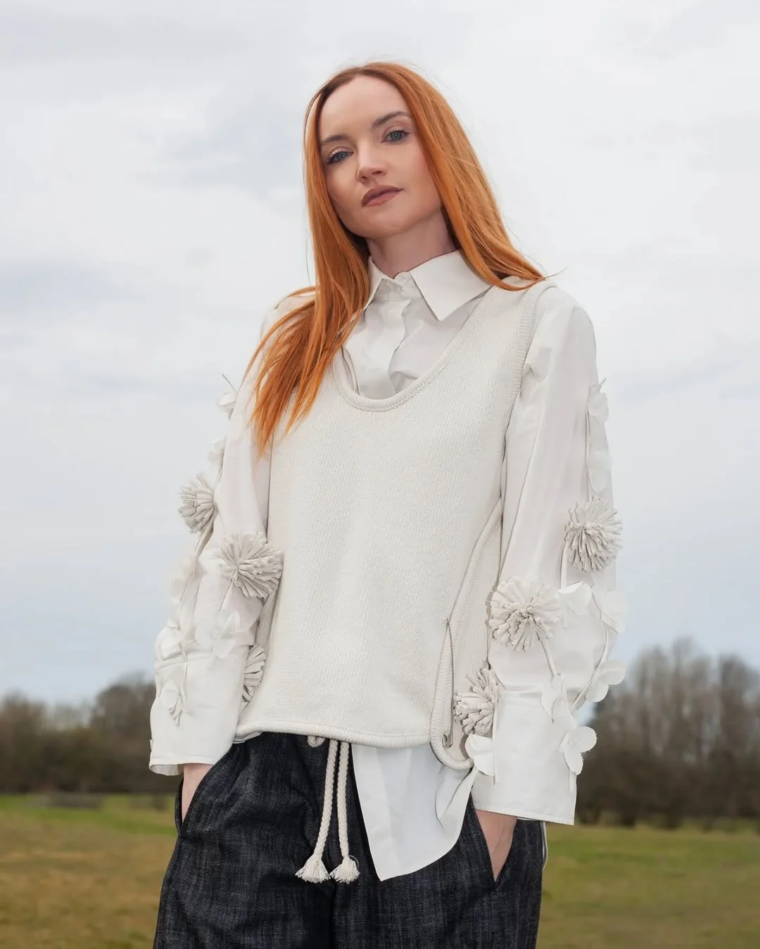 A woman with red hair stands outdoors on a cloudy day, wearing a white collared shirt, a beige vest with 3D floral details on the sleeves, and dark drawstring pants.