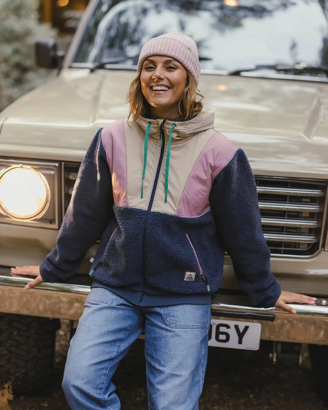 A young woman smiling and leaning against a beige vintage car with round headlights, wearing a pink beanie, a color-blocked windbreaker, and jeans, outdoors.
