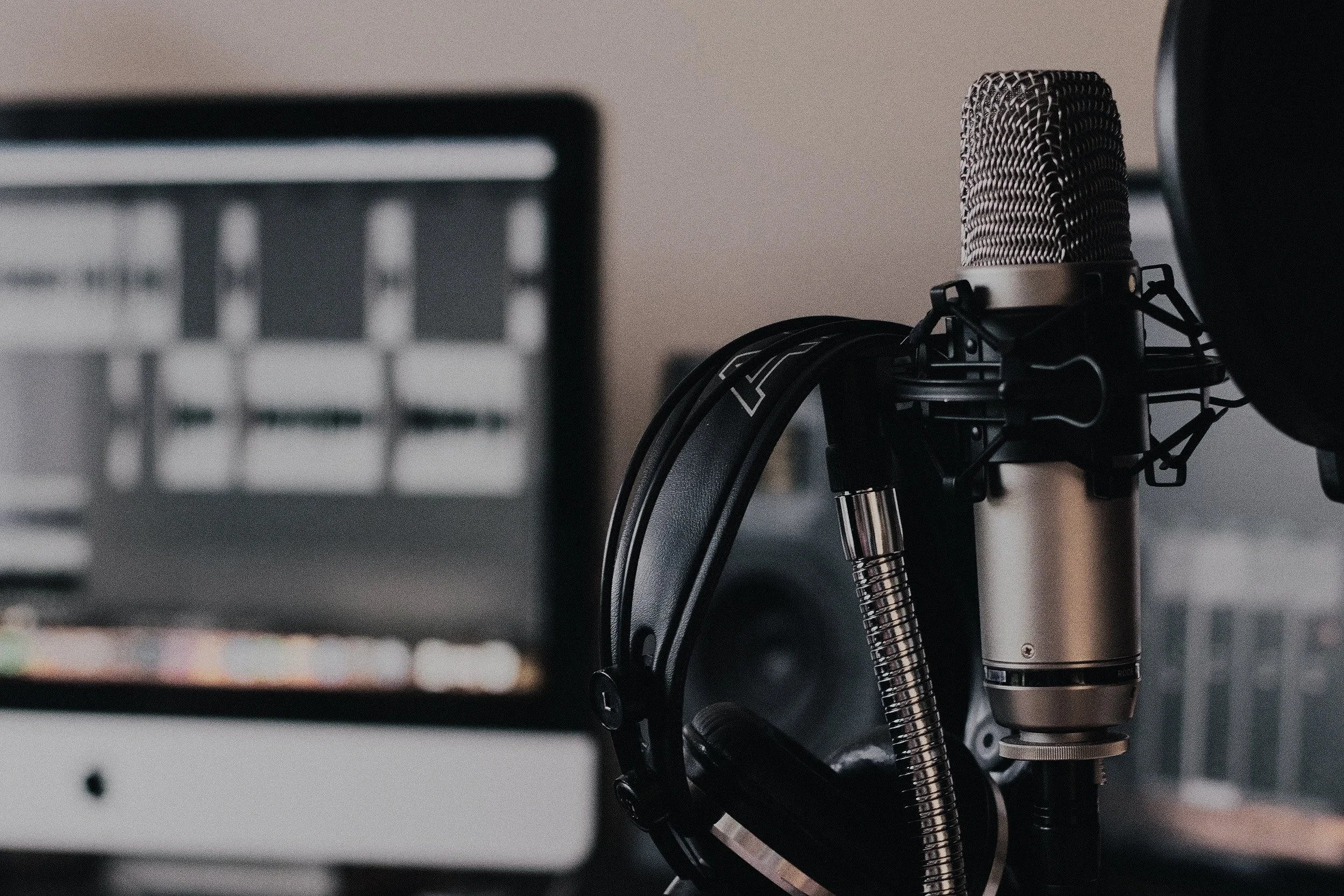 Close-up of a professional microphone mounted on a shock mount and a pop filter in a recording studio. Out of focus in the foreground is a piece of audio equipment or computer screen.