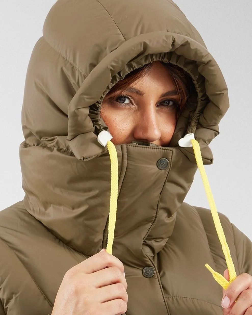Person wearing a tan puffer jacket with a hood pulled tight around their face, holding yellow drawstrings near their chin.