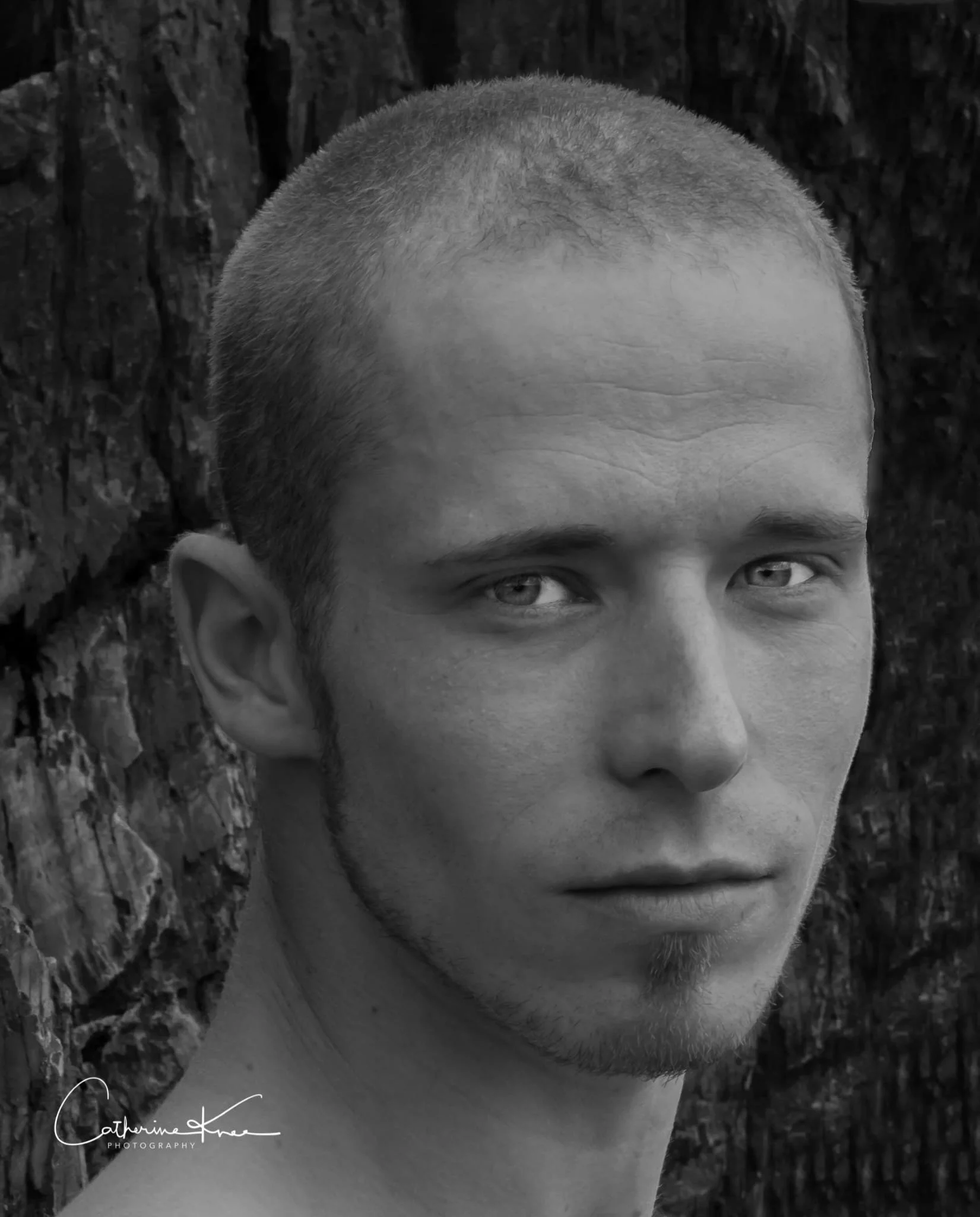 A black and white portrait of a young man with a closely shaved head, looking slightly to his left, with a blurred tree bark background.