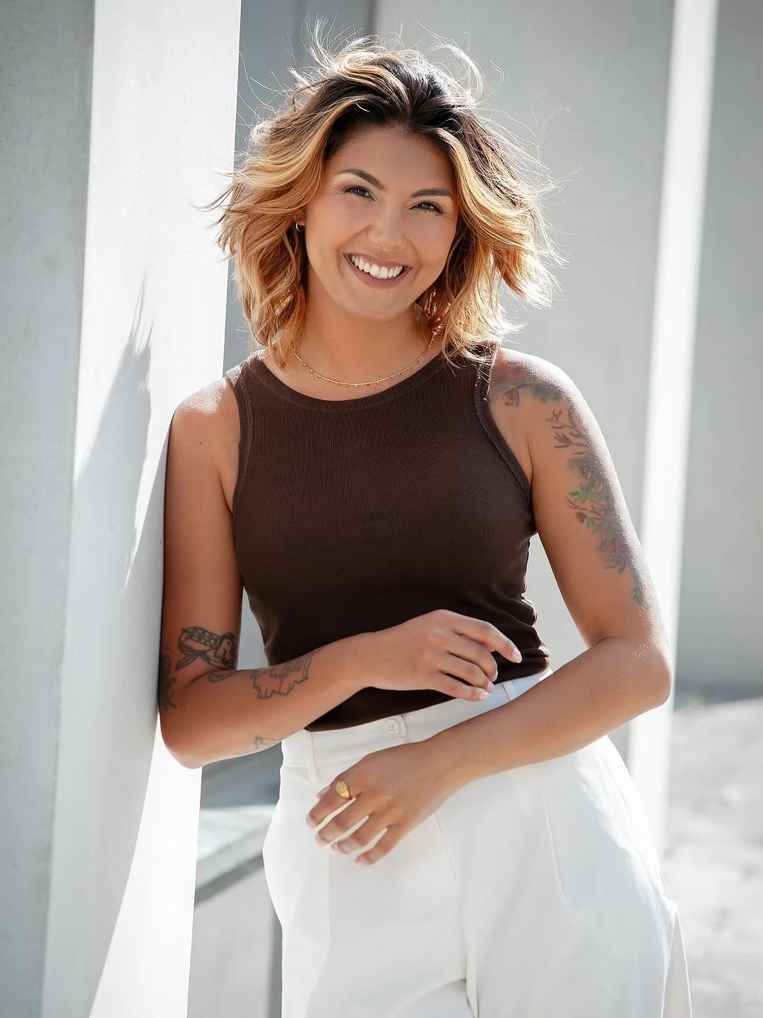 A woman with wavy blonde hair, wearing a dark sleeveless top and white high-waisted pants, smiling and leaning against a white wall in bright sunlight.