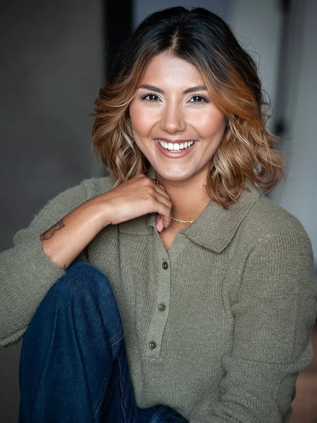 A woman with shoulder-length wavy hair, smiling, wearing a green button-up sweater and blue jeans, sitting indoors with a neutral background.