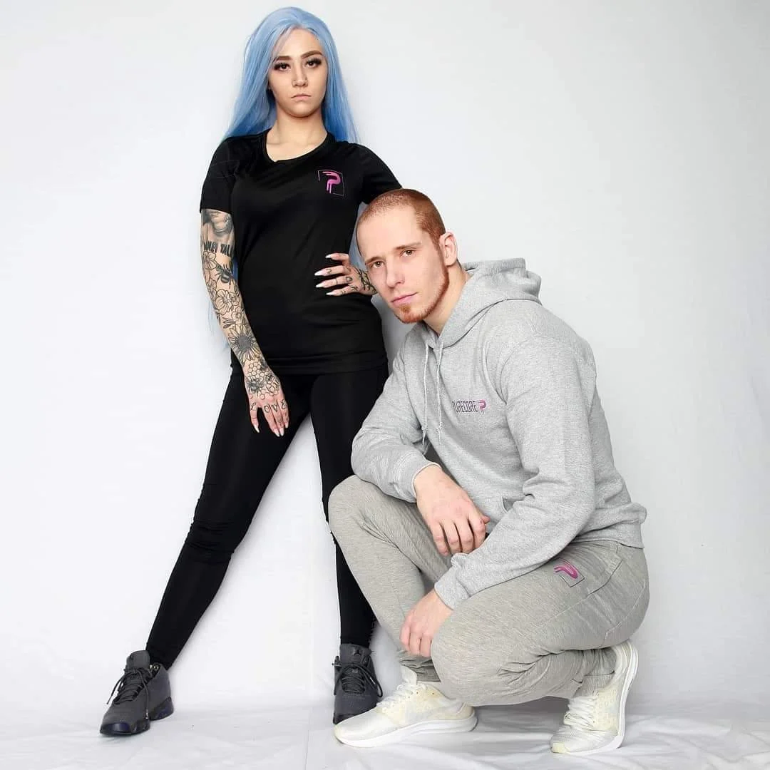 A woman with long, light blue hair and tattoos on her arms wearing a black sports outfit, and a man with short hair in a gray hoodie and sweatpants, pose against a plain white background.