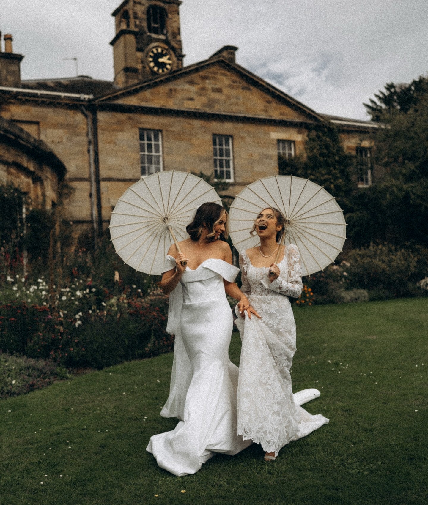 Two women in wedding dresses holding umbrellas and laughing outdoors in front of a stone building with a clock tower.