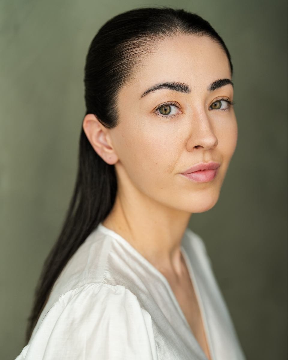 A young woman with dark hair, light skin, and hazel eyes smiling softly, wearing a white blouse, against a neutral green background