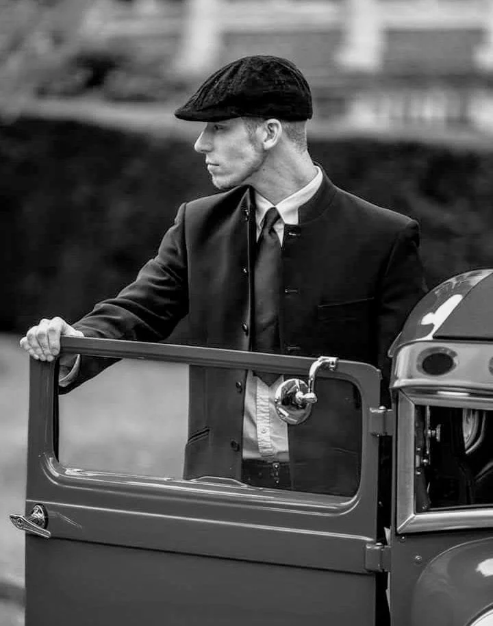 A man dressed in vintage attire with a flat cap and suit, standing next to an open car door in black and white.