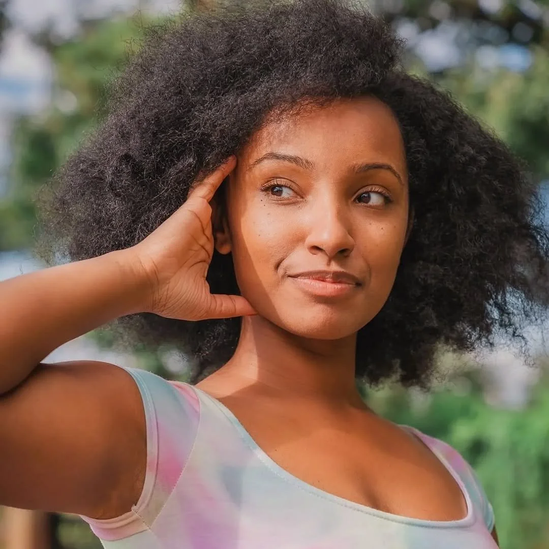 A woman with curly black hair, wearing a pastel tie-dye shirt, outdoors with blurred trees in the background, looking to the side with a gentle smile and resting her hand on her cheek.