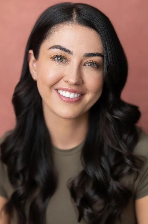 A woman with long, wavy black hair smiling against a pink background.