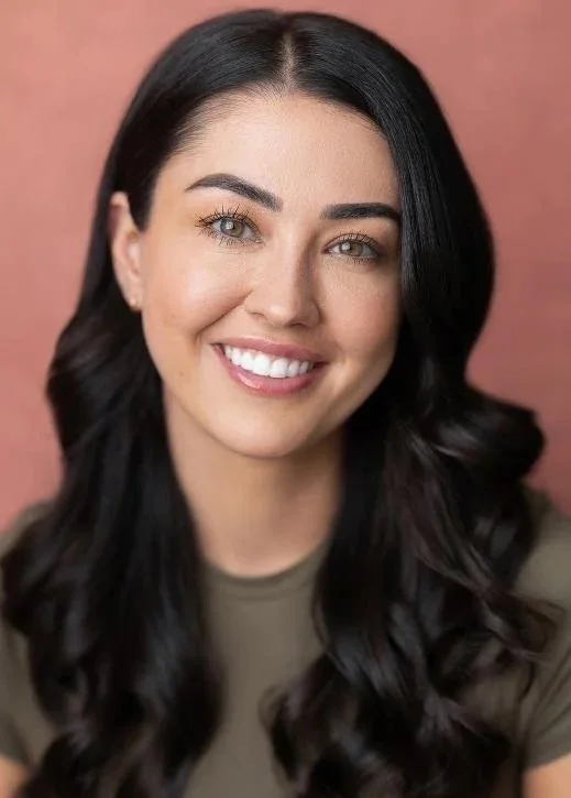 Portrait of a woman with long black wavy hair, blue eyes, and a bright smile, against a pinkish background.