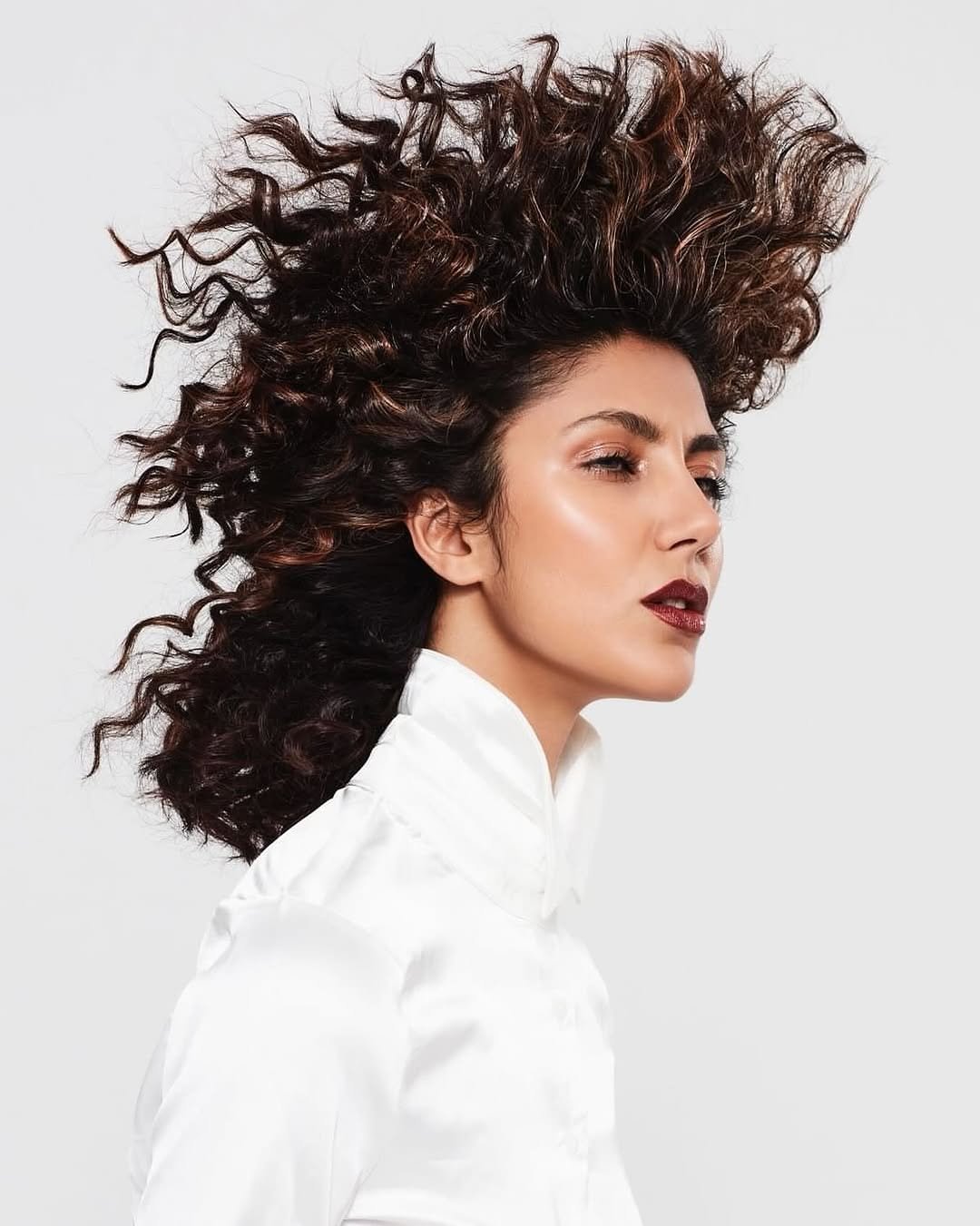 A woman with voluminous, curly, dark brown hair with lighter highlights, wearing a white high-neck satin blouse, against a plain white background.