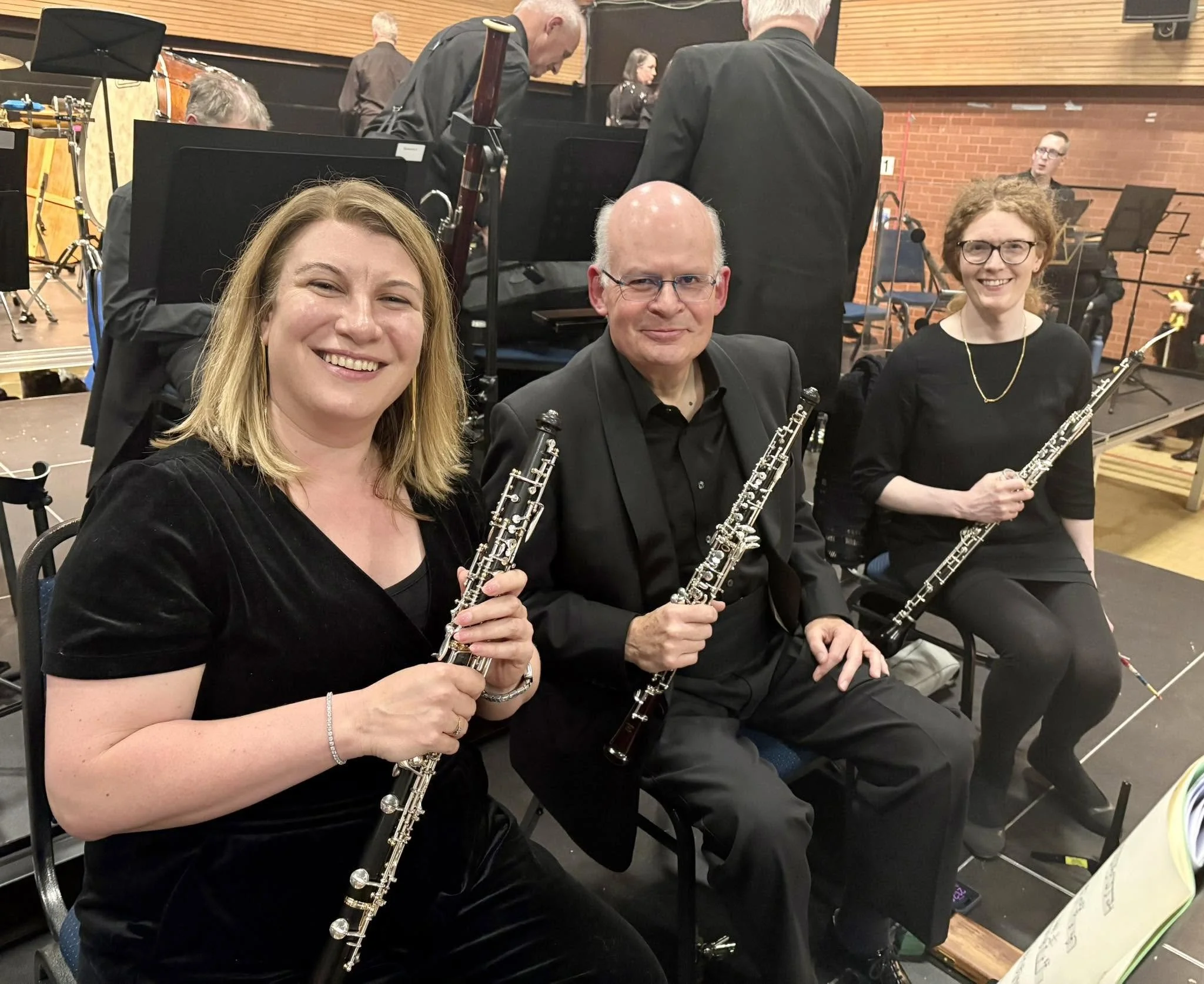 Three musicians seated in a row holding clarinets, with a music stand and an orchestra behind them in a concert hall setting.