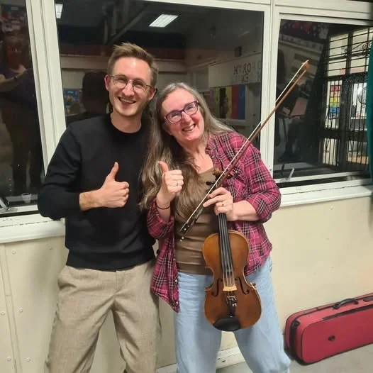 A young man and a woman holding a violin, smiling and giving a thumbs-up.