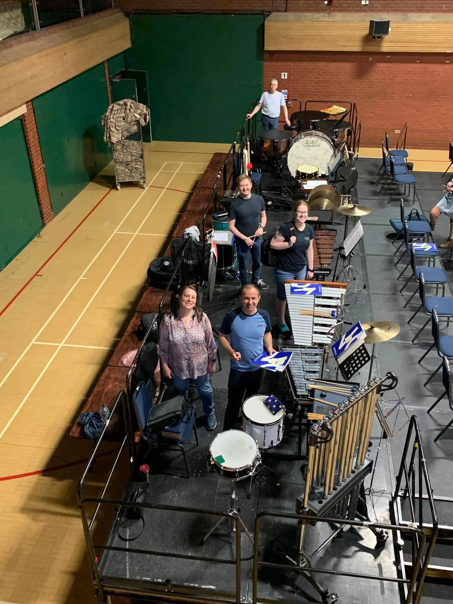 A group of people in a gymnasium setting with music and percussion instruments set up, including drums, xylophones, and cymbals, preparing for a performance or rehearsal.