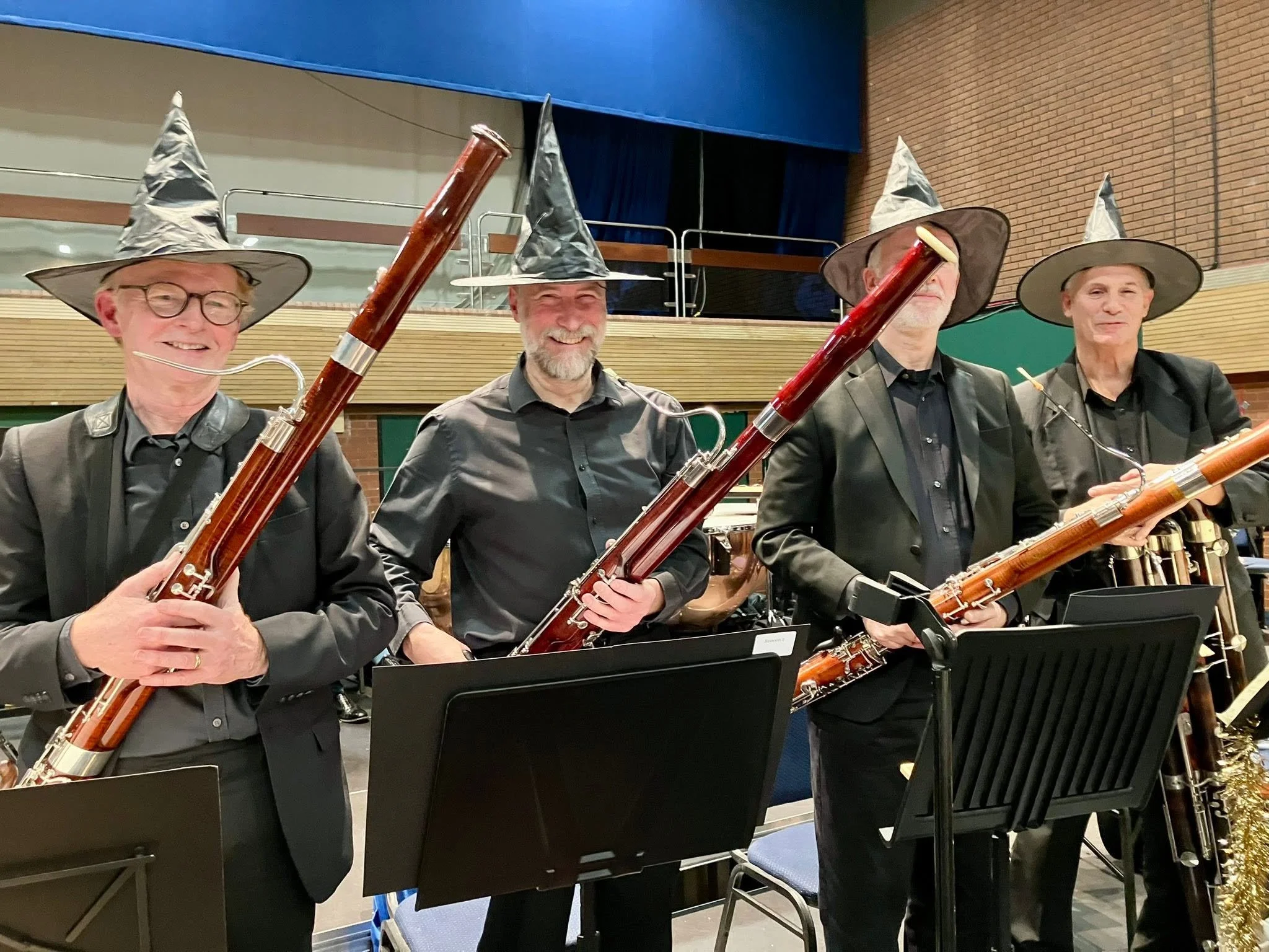 Four musicians in black suits and wizard hats playing bassoons in an indoor concert venue.