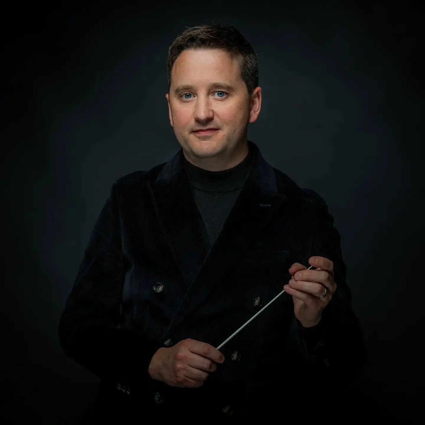 A man with short brown hair and blue eyes dressed in a black jacket and black turtleneck holding a conductor's baton against a dark background.