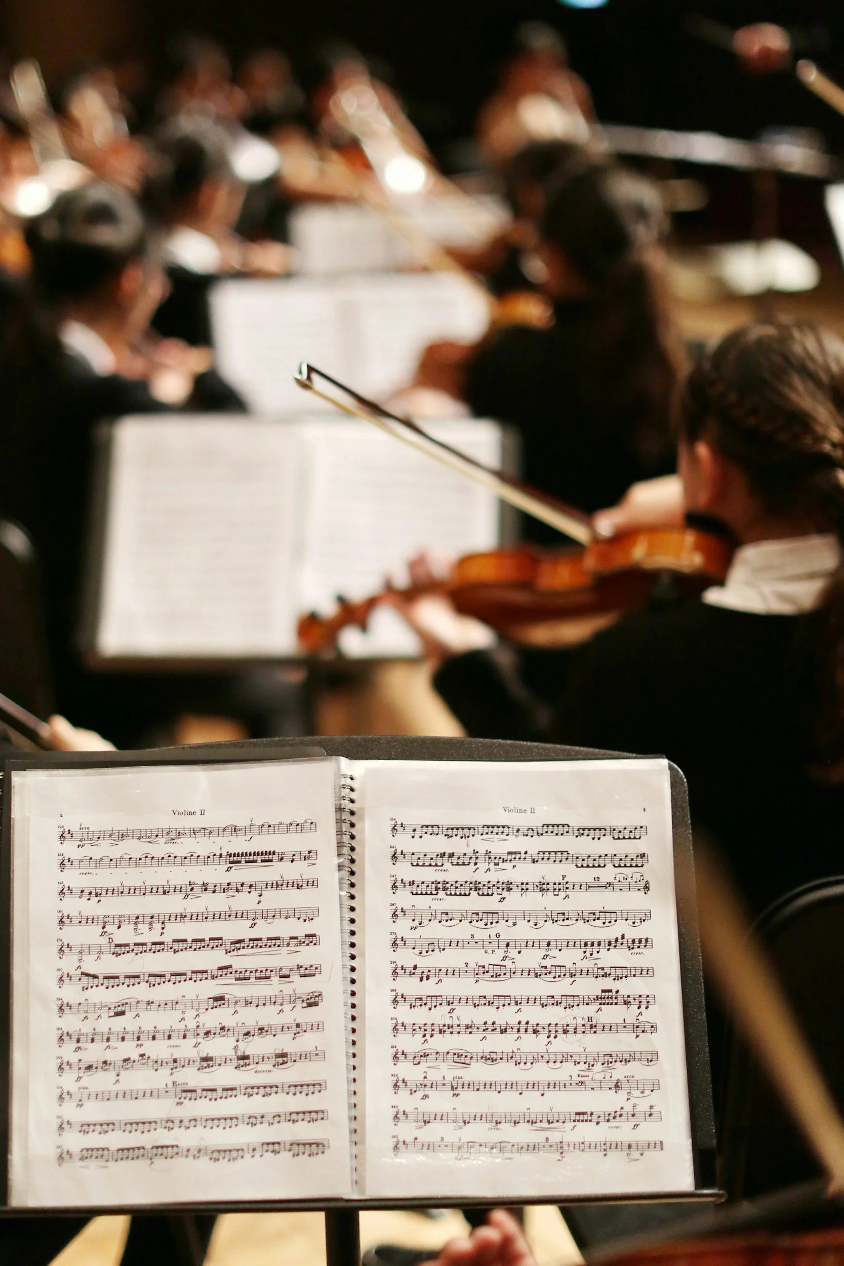 Music score with notes and markings open on sheet music stand, with an orchestra and conductor in the background.