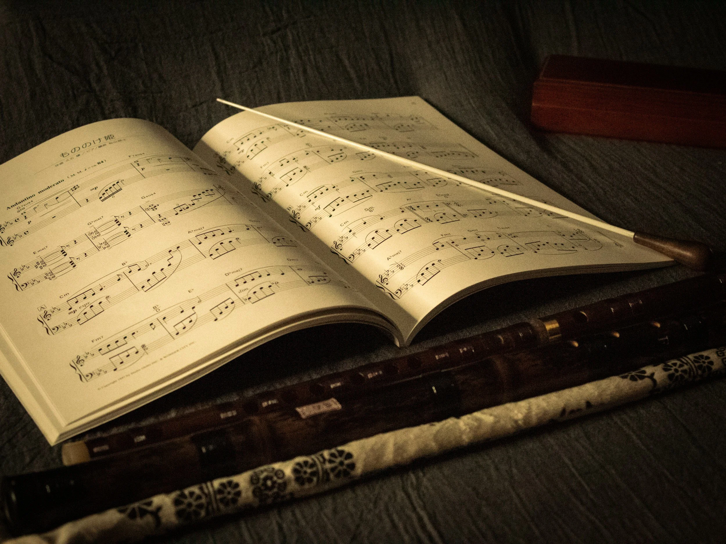 Open sheet music book with musical notes and titles, resting on a dark textured surface, with traditional wooden and decorative items nearby.