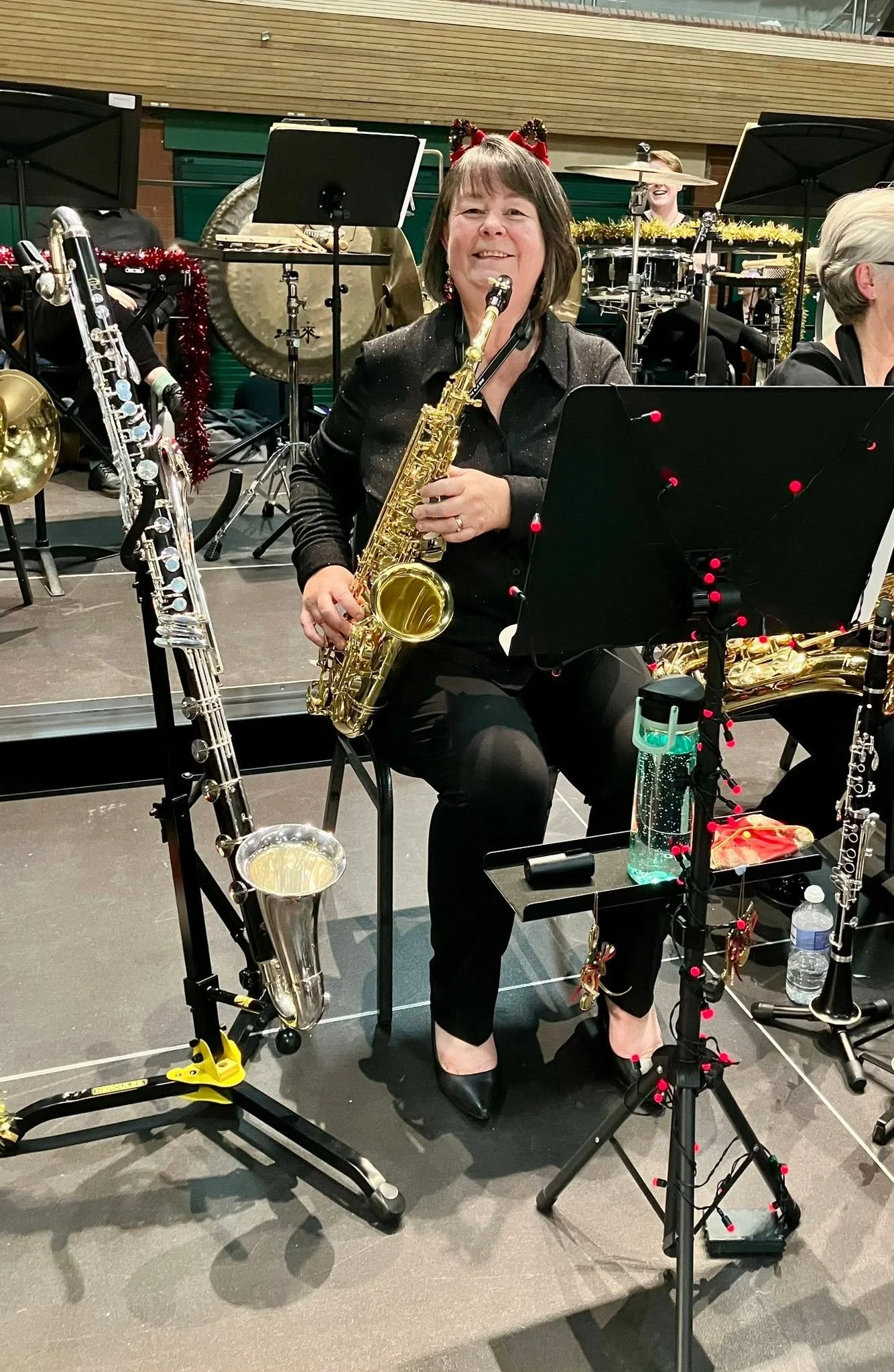 Woman holding a saxophone in a band performance setting, surrounded by musical instruments, music stands, and Christmas decorations.