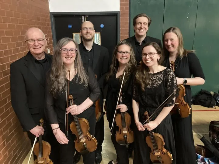 Group of seven musicians, five women and two men, holding violins, posing for a photo backstage in a school or community center hall, dressed in black concert attire.