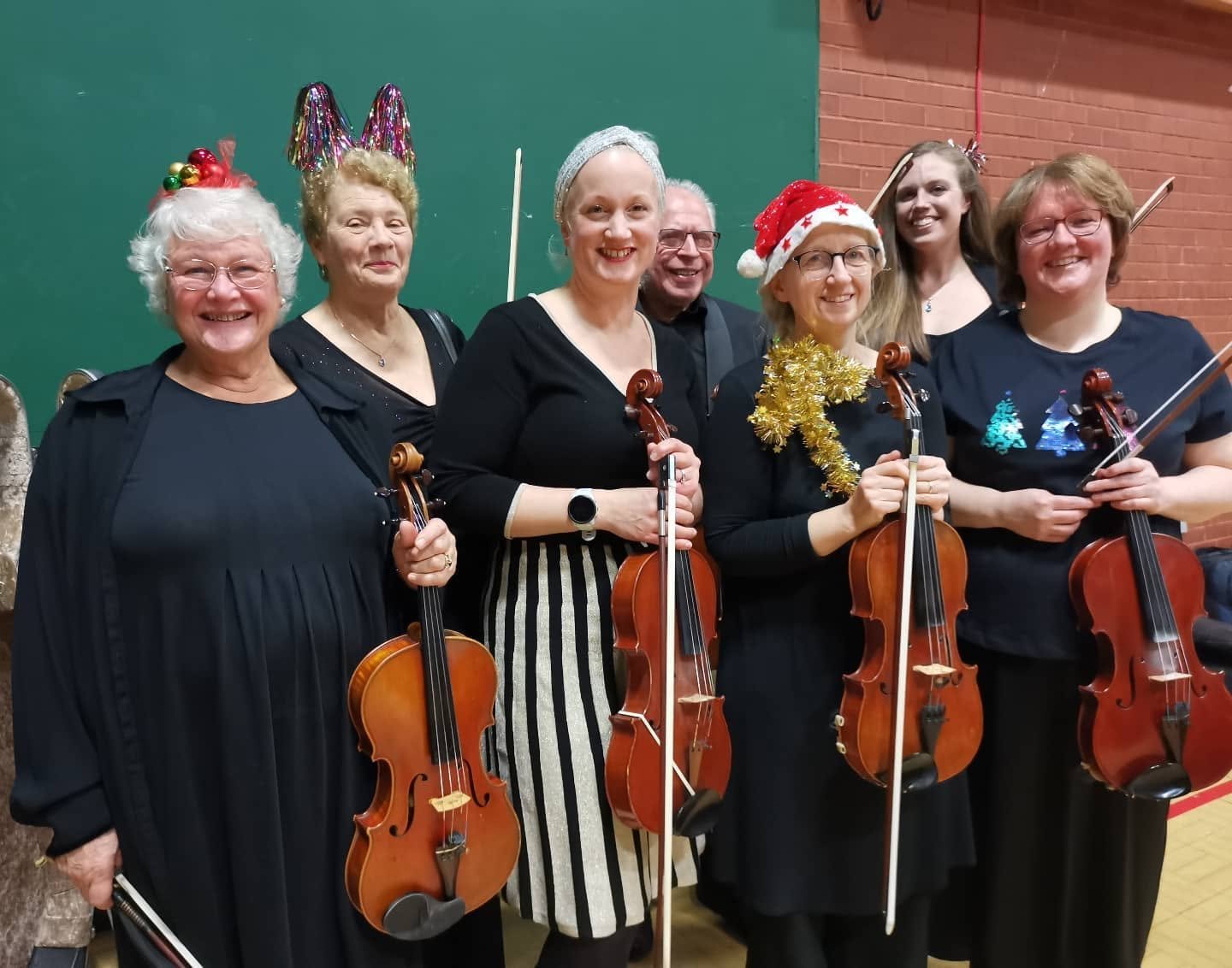 Group of seven people smiling, holding violins, dressed festively with holiday accessories, in front of a green and brick wall.