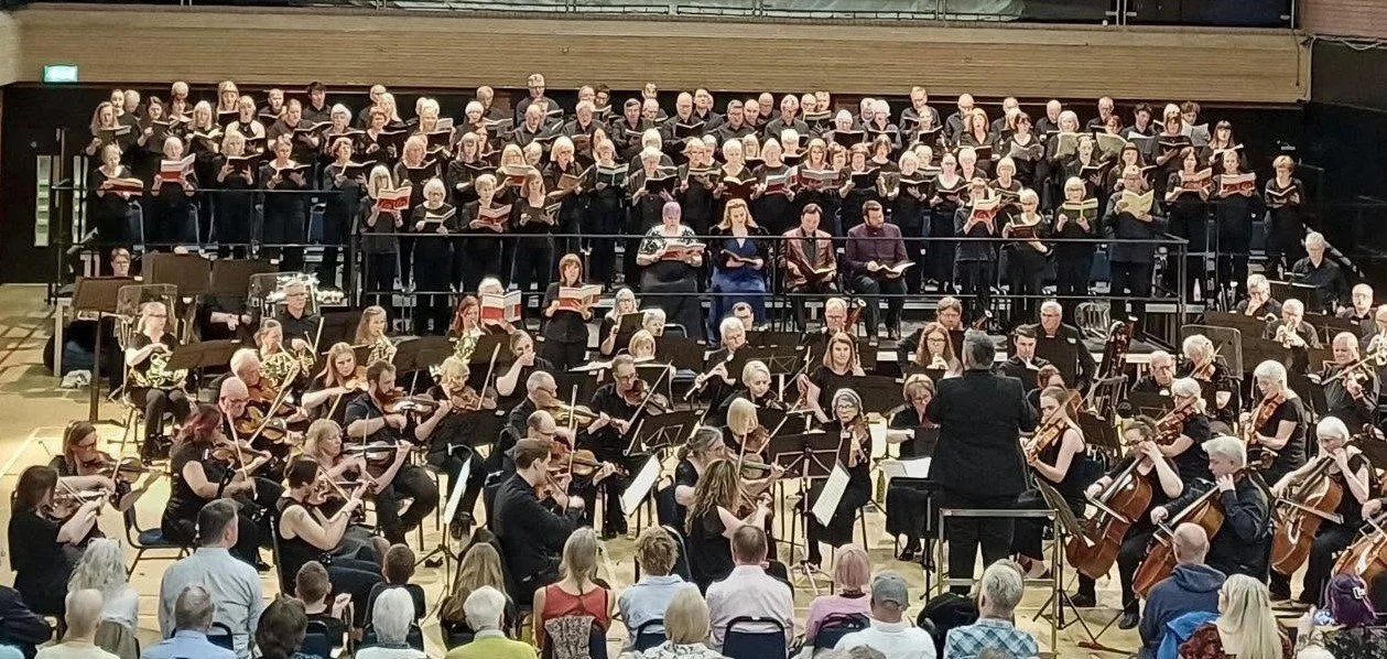 Large orchestra and choir performing in a concert hall with an audience watching.
