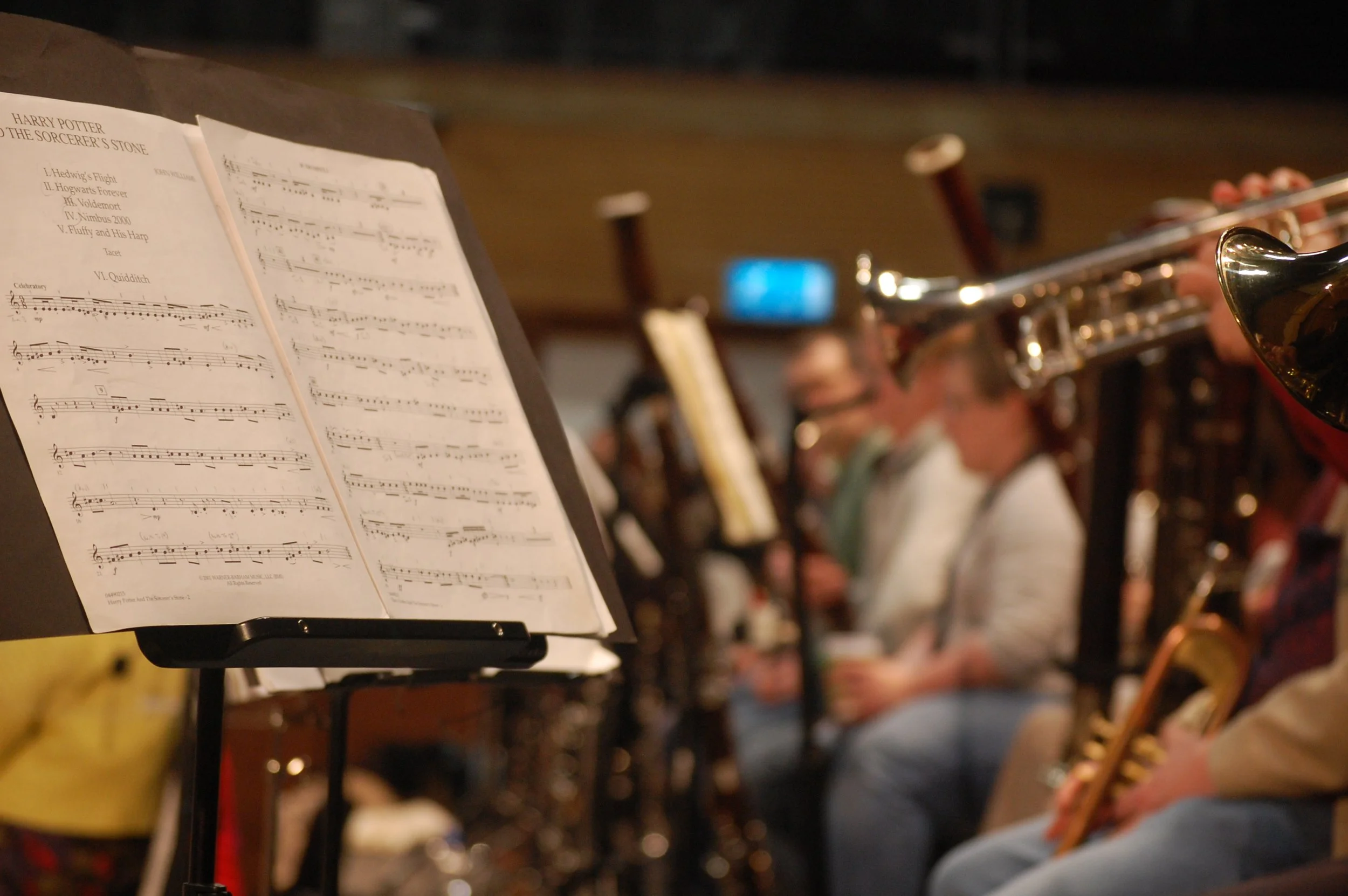 A music stand holding sheet music titled 'Harry Potter and the Sorcerer's Stone,' with musicians playing instruments in the background, including a person playing a flute.