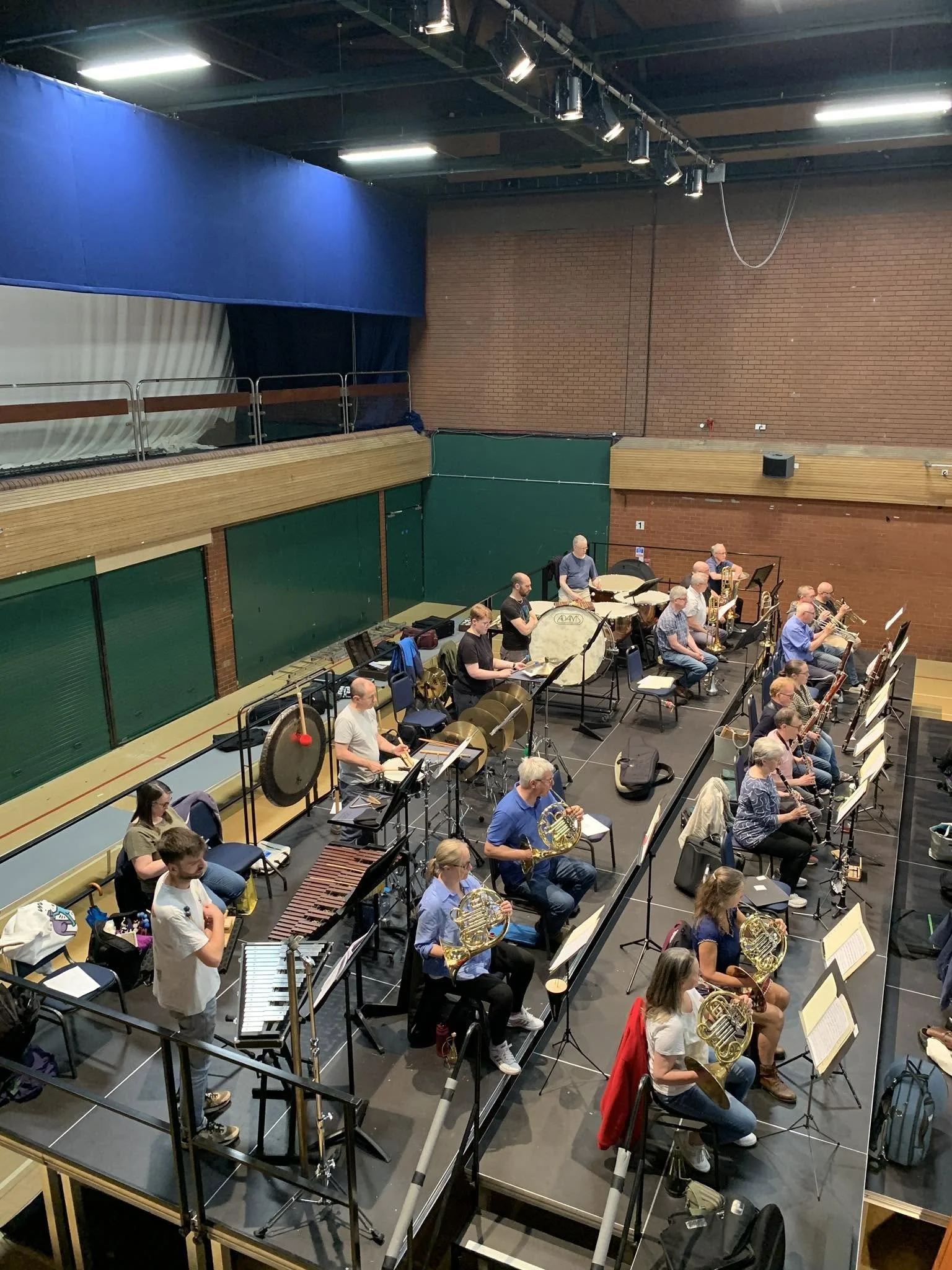 An orchestra rehearsal or performance taking place in a large auditorium. Musicians are seated with various instruments, including French horns, percussion, and brass, while a conductor stands at the front.