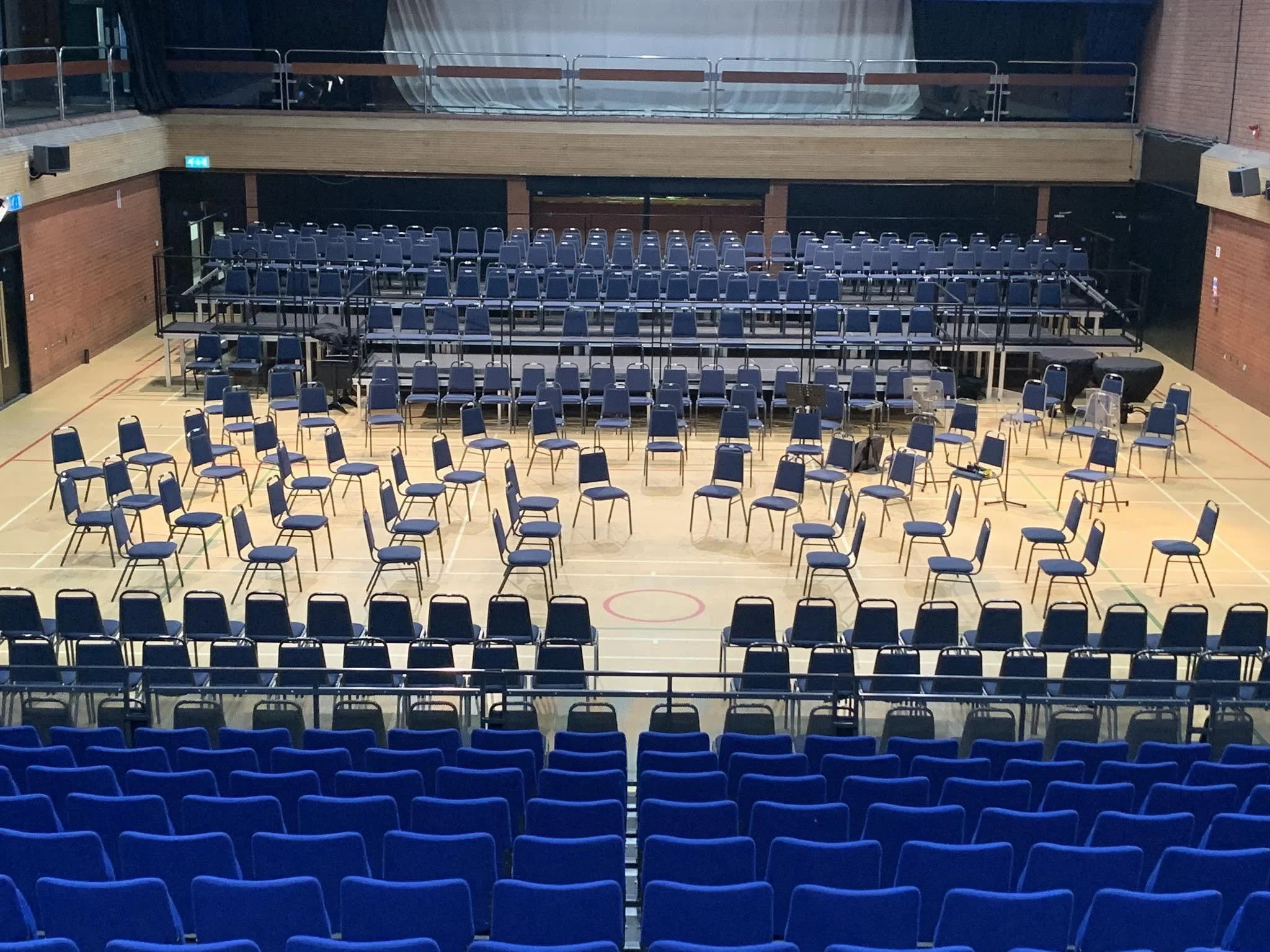 Indoor auditorium with multiple rows of blue chairs arranged in front of a stage with a grand piano. The space has brick walls, a wooden balcony, and a high ceiling with stage lighting.