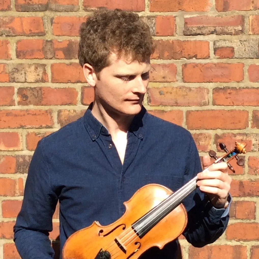 A young man with curly brown hair holding a violin, standing against a brick wall, looking down at the violin.
