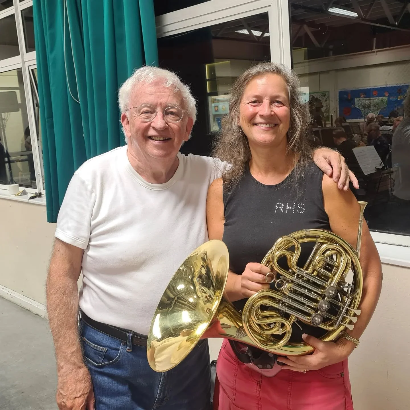 Two smiling people, a man with glasses in a white T-shirt and a woman holding a French horn and wearing a black sleeveless top with 'RHS' written on it, inside a room with turquoise curtains and large windows.